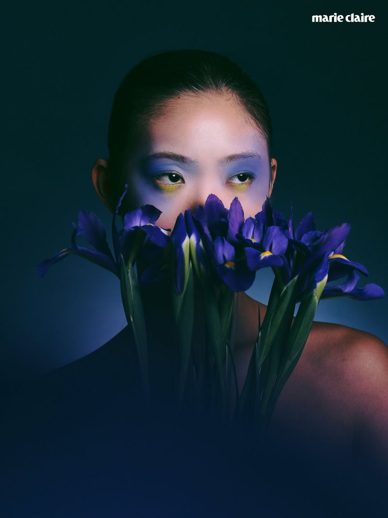 Close-up portrait of a woman with vibrant blue eye makeup holding purple irises covering her lower face against a dark studio background