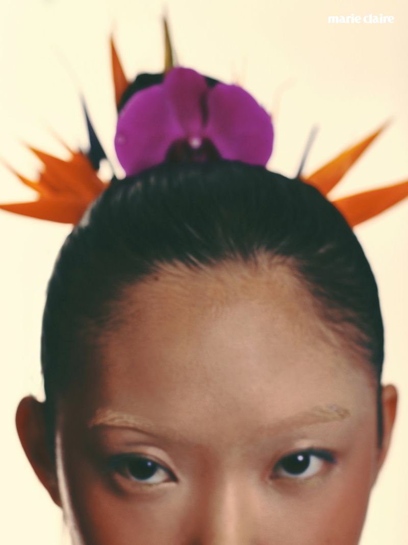 Close-up portrait of a woman with dark hair styled up, adorned with a vibrant purple flower and orange bird-of-paradise petals against a light background.