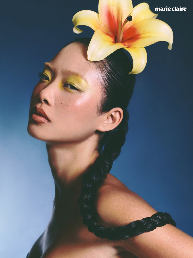 Model with yellow eye shadow and a large yellow lily in her braided dark hair against a gradient blue studio backdrop