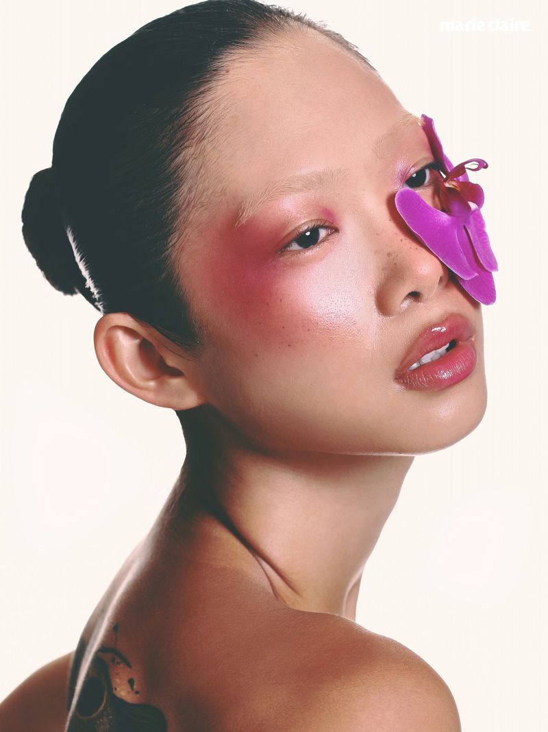Close-up of an Asian woman with a pink orchid over her eye. The image features a beauty and fashion theme with soft lighting.