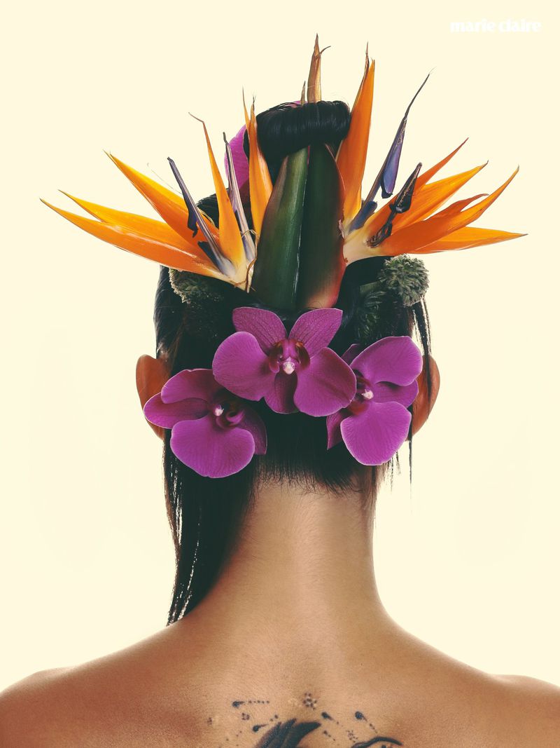 Rear view of a person with elaborate floral hair styling featuring bird of paradise and purple orchids against a cream background.