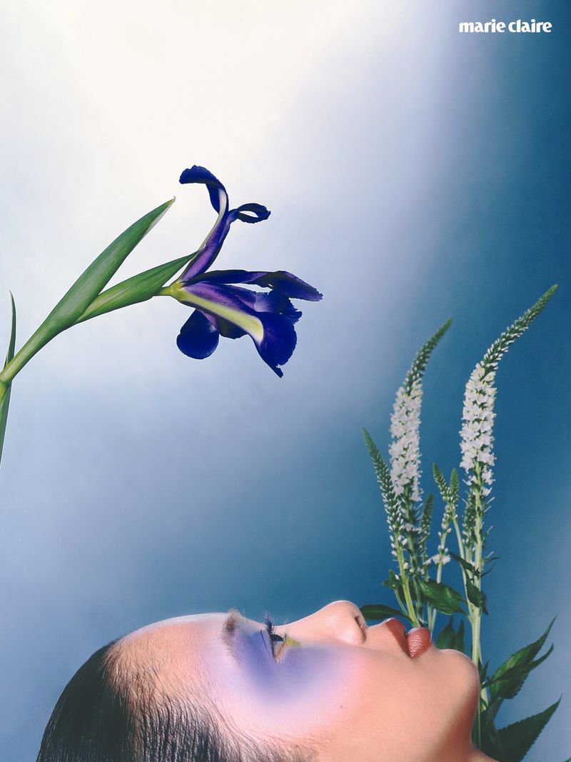 Close-up profile of a woman with purple eye makeup alongside a dark blue iris flower and white spiky floral stems