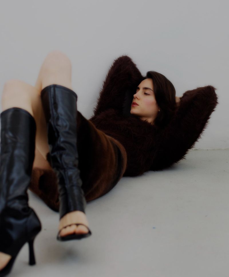 A woman in a brown, furry sweater and black boots lies on a gray floor with her hands behind her head, eyes closed, in a studio setting.