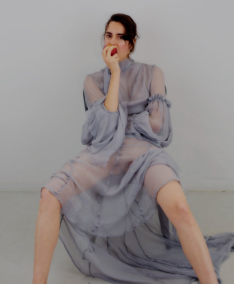 Model seated on a chair wearing a sheer grey dress holding an apple in front of her face against a white background