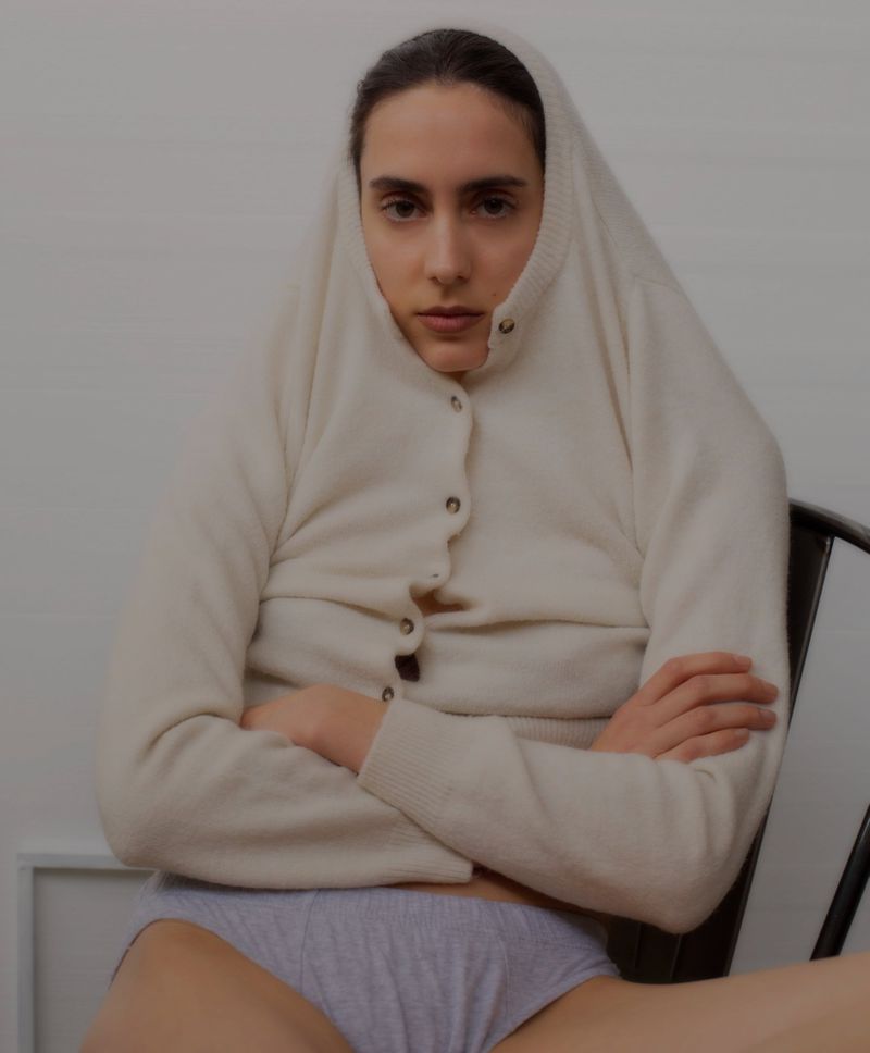 Model wearing a cream cardigan over her head and arms crossed while seated against a plain background
