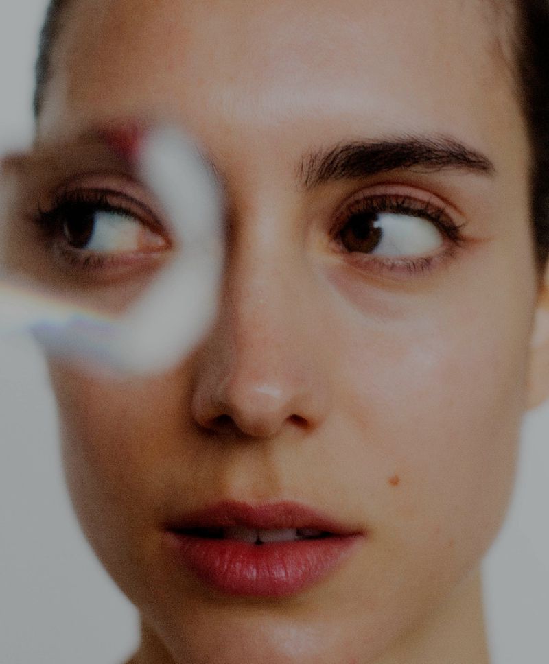 Close-up portrait of a woman with natural makeup, looking sideways behind a curved frosted glass element.