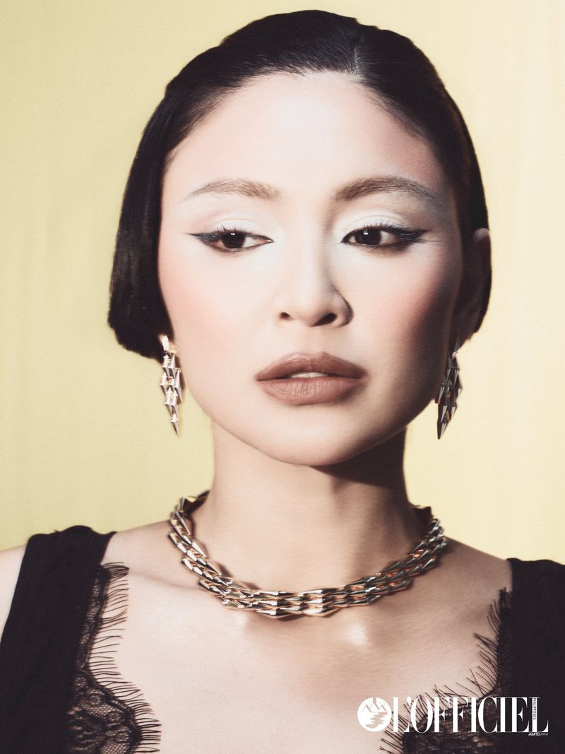 Close-up portrait of a woman with dark hair, winged eyeliner, silver chain necklace, and drop earrings against a pale yellow background.