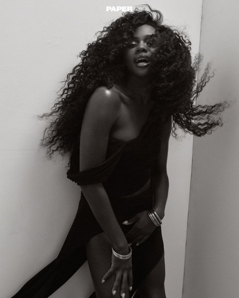 Black and white portrait of a woman with voluminous curly hair wearing a dark one-shoulder top and stacked bangles against a plain corner wall.