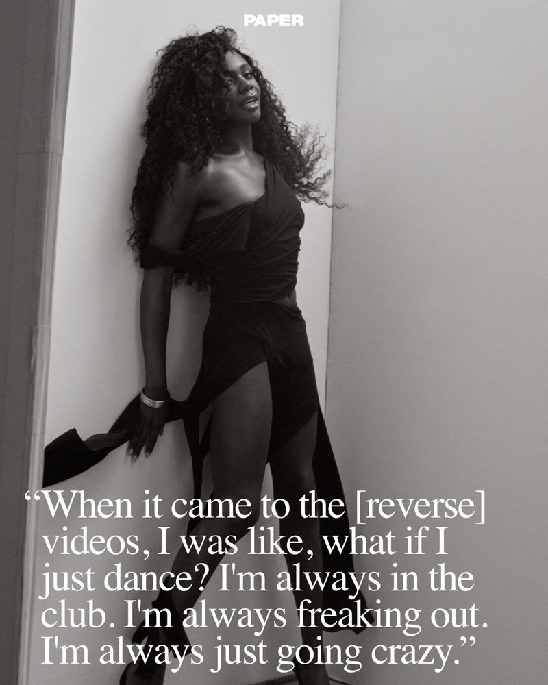 Monochrome portrait of a woman with long dark curly hair, in an asymmetrical black dress, leaning against a wall in a studio setting.