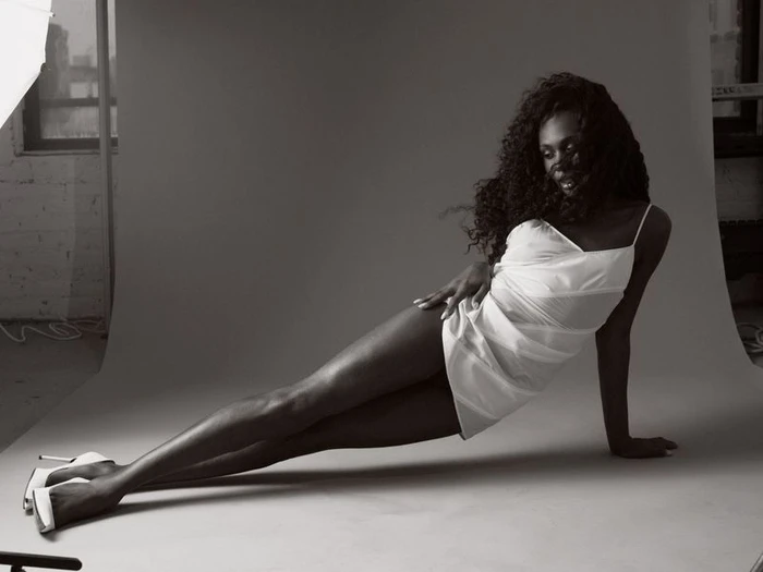 Black woman in white slip dress and high heels posing on a grey seamless studio backdrop for Paper Magazine.