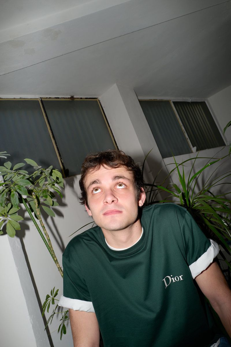 Young man in a dark green t-shirt with rolled white cuffs looking upward indoors with plants flanking him.