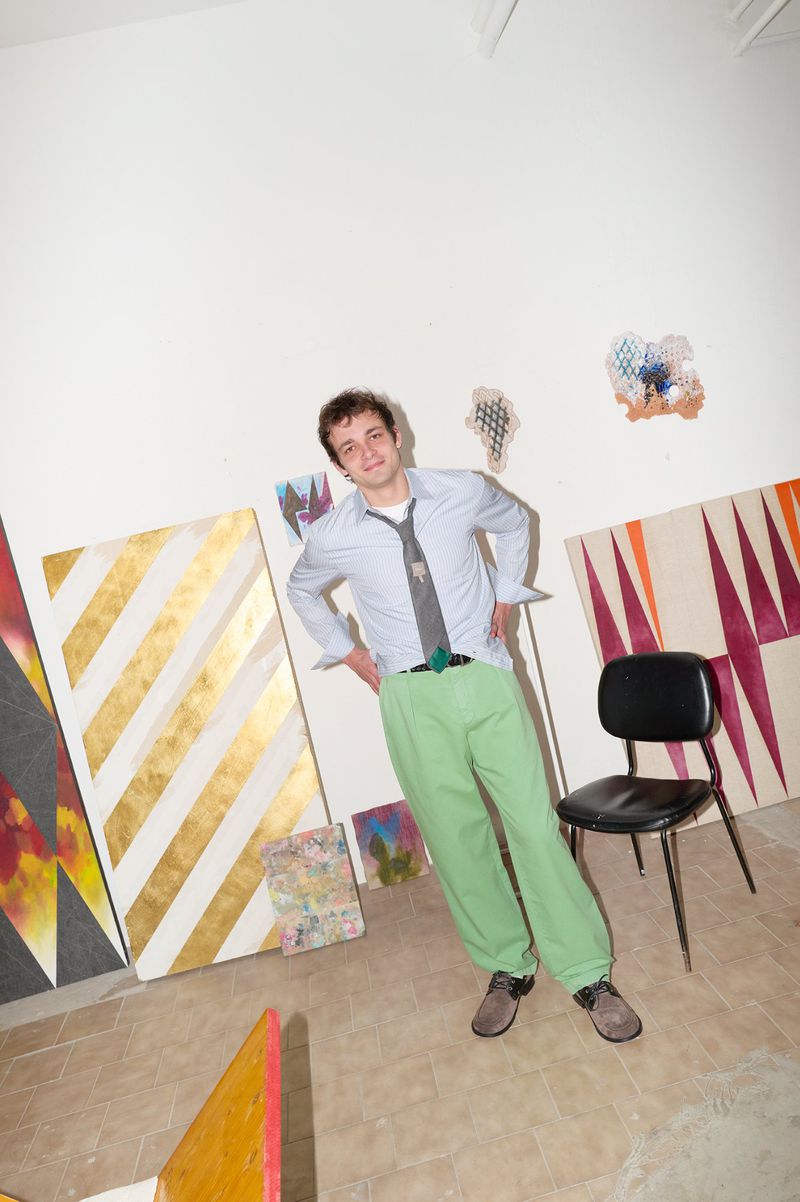 Young man in light blue shirt and green trousers standing in an art studio with paintings and a chair.