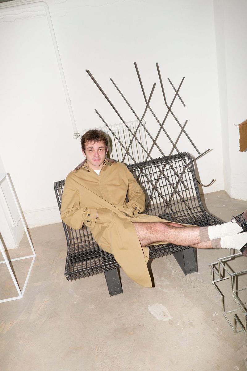 Young man in a tan trench coat seated on a wire mesh bench inside a white room