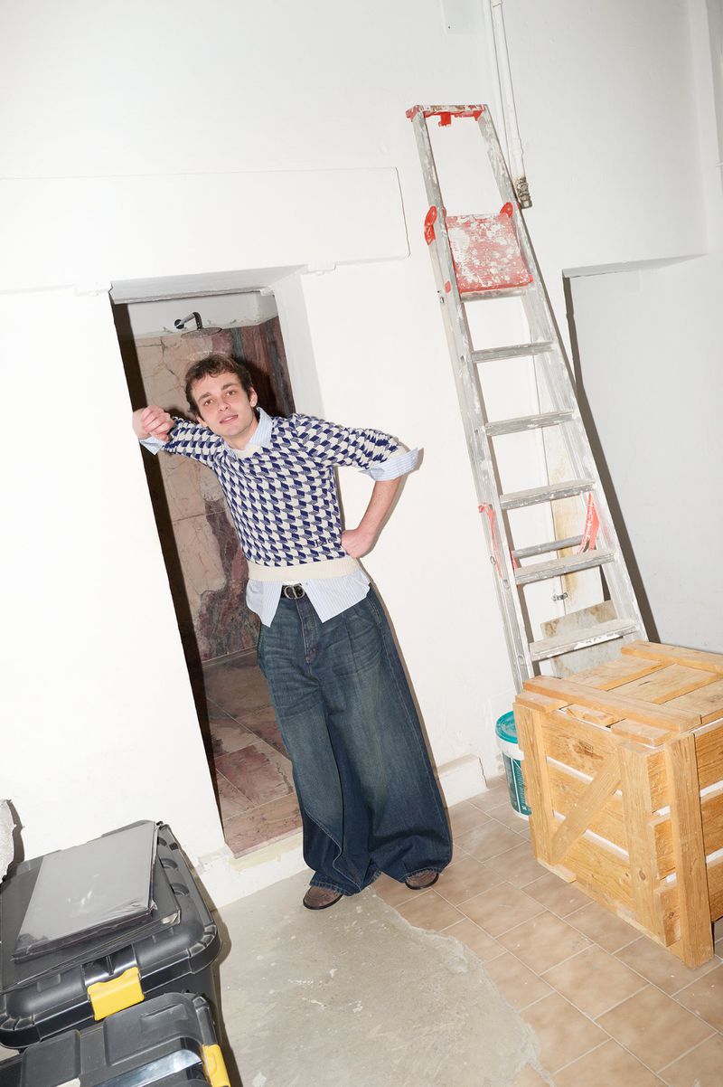 Young man poses indoors wearing baggy jeans and a patterned top, with a ladder, tool boxes, and unfinished walls in the background.