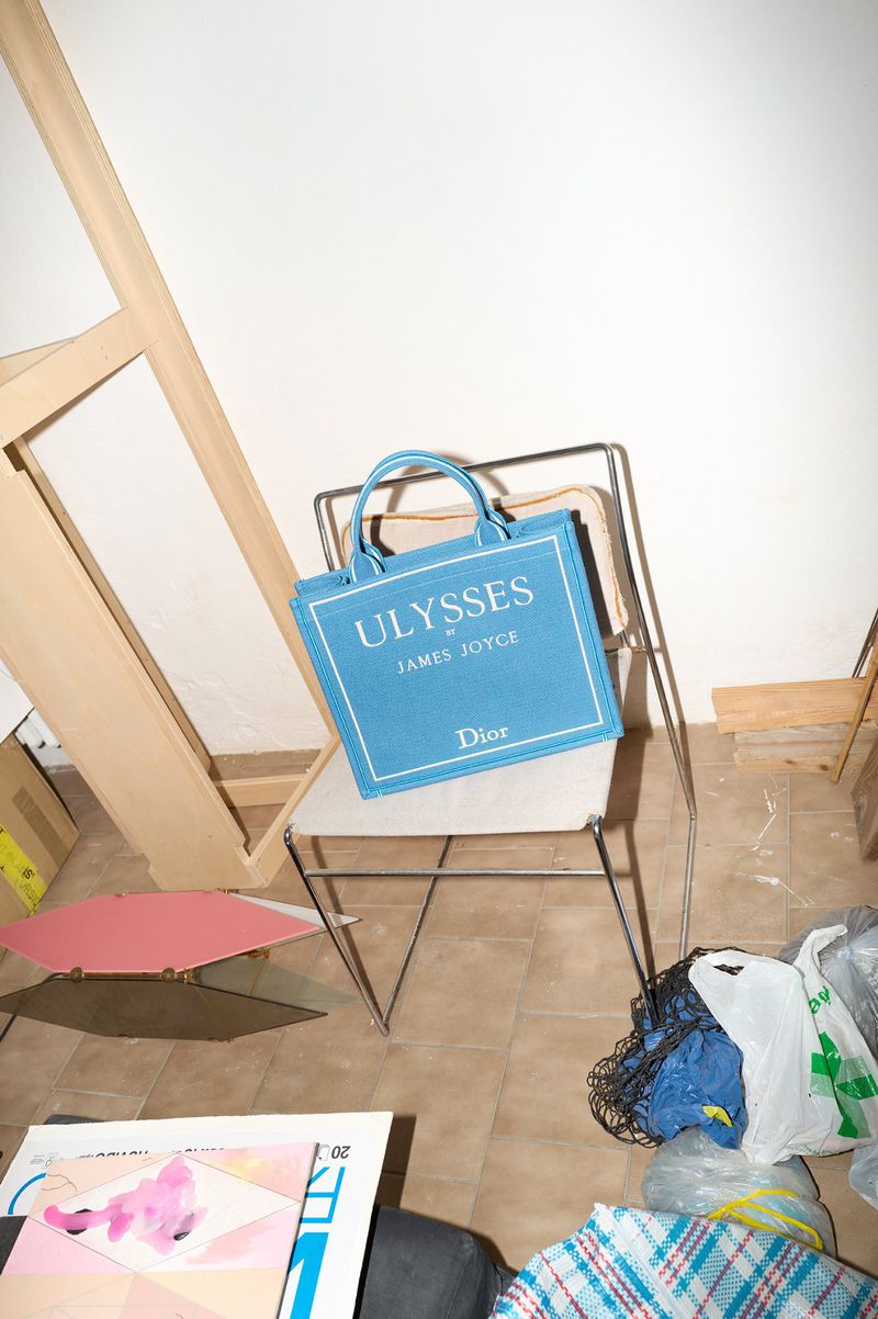 A blue Dior tote bag resting on a modern metal-frame chair in an indoor room with tiled floors and various objects.