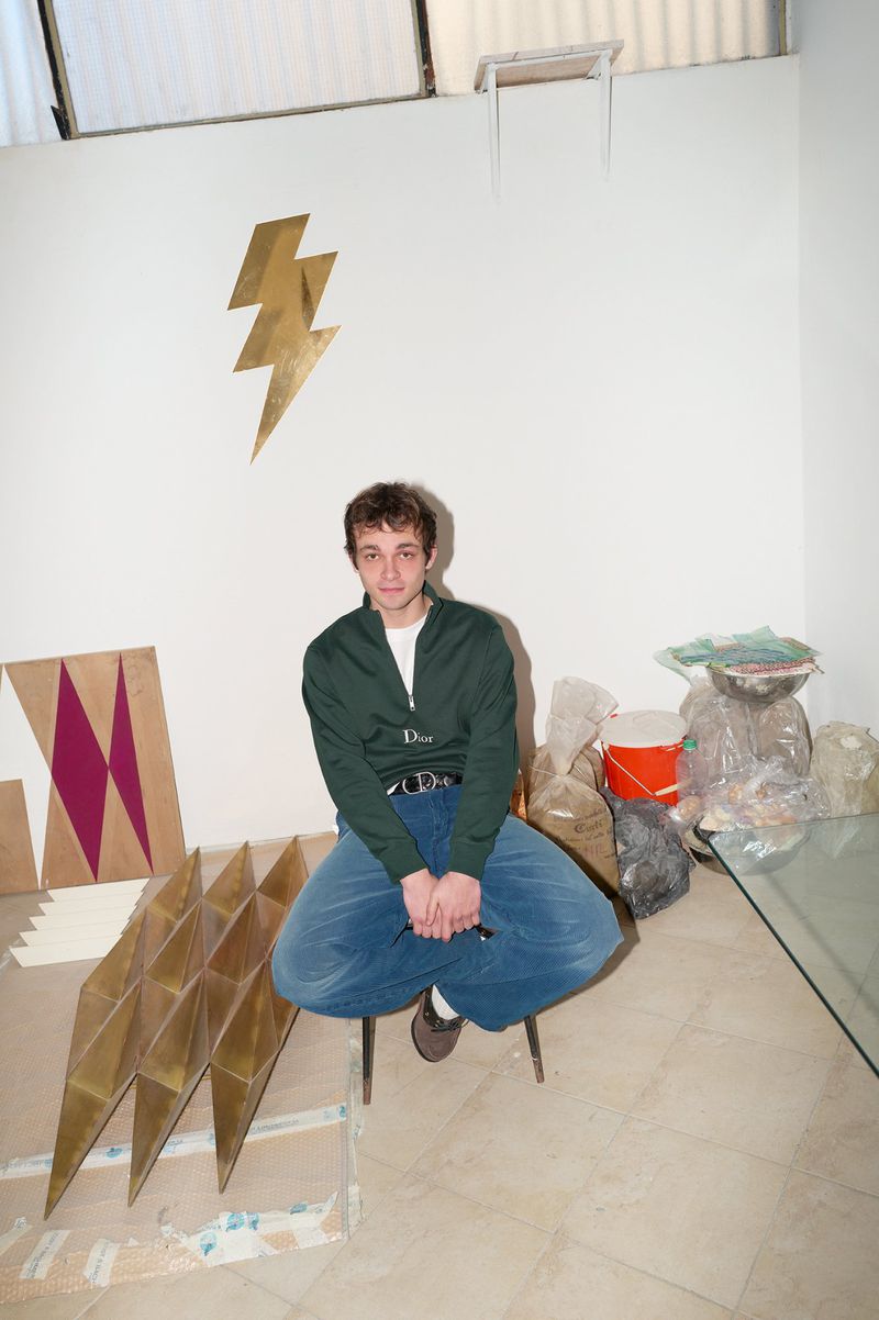 Young man in a green zip sweater and blue denim pants seated cross-legged indoors near metallic sculptures.