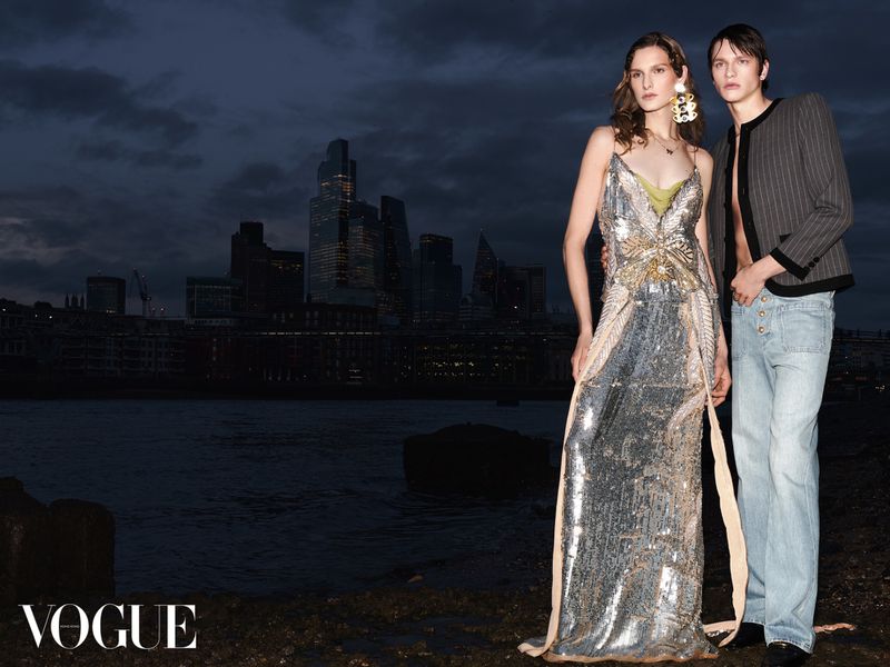A female model in a sparkling silver gown and a male model in a striped jacket pose outdoors against a city skyline at dusk.
