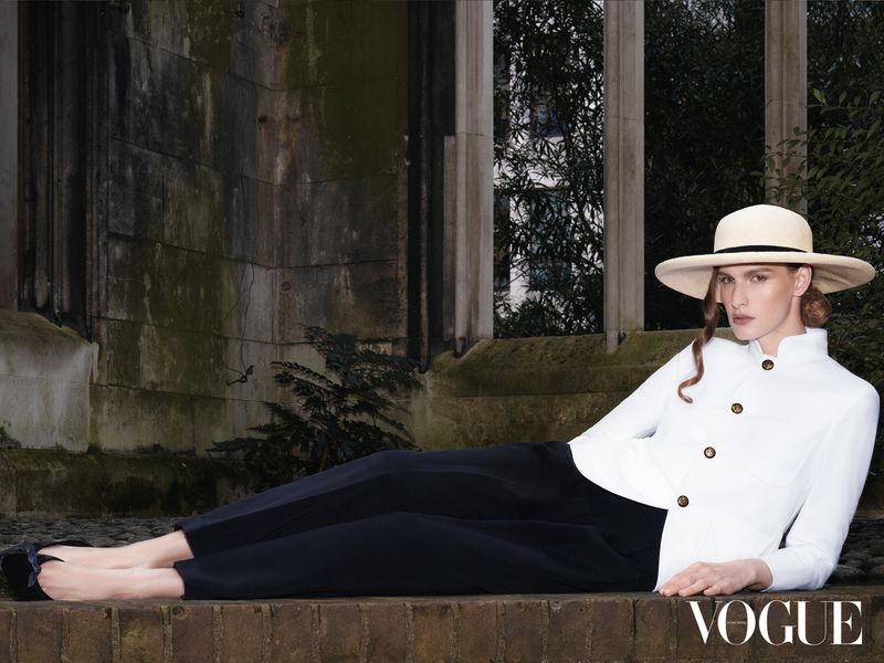 Model in wide-brim hat, white jacket and dark trousers reclining outdoors near stone architectural columns