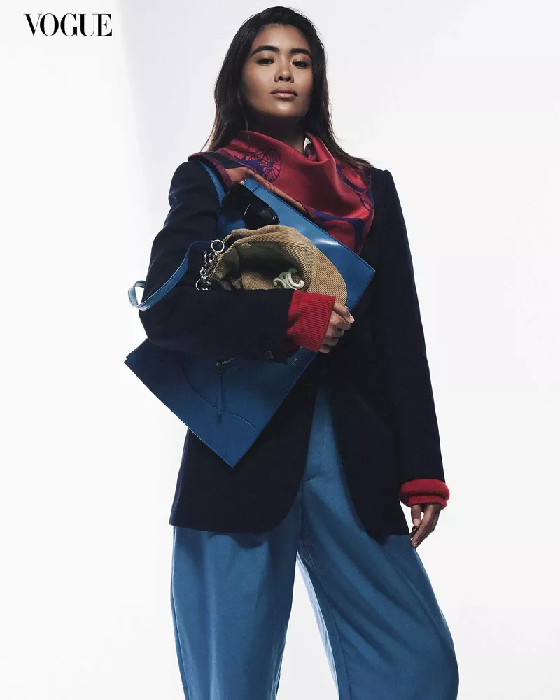 Woman wearing dark blazer, red patterned scarf, and blue trousers holding a blue bag and tan cap against white seamless background.