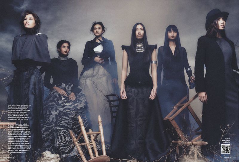 Group of six female models in dark avant-garde fashion standing outdoors amid scattered wooden chair frames.