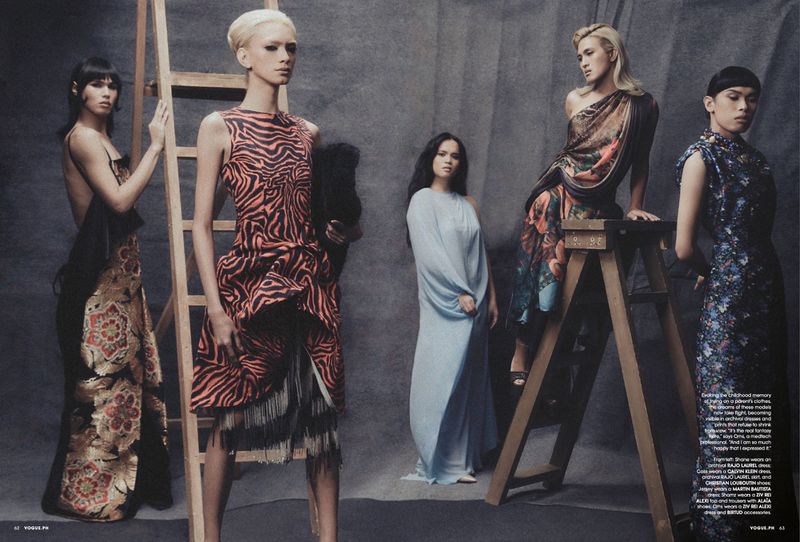 Group of five models in stylized designer dresses posing together against a textured grey studio backdrop with wooden ladders.
