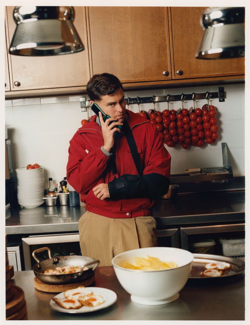 Man wearing a red jacket and beige trousers talking on a mobile phone inside a commercial kitchen