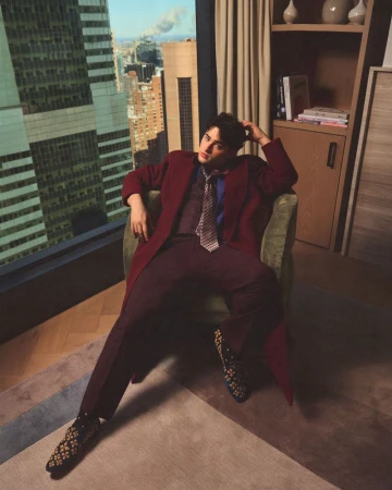 Young man in a burgundy coat and patterned tie seated in an armchair by a tall window overlooking a city skyline.