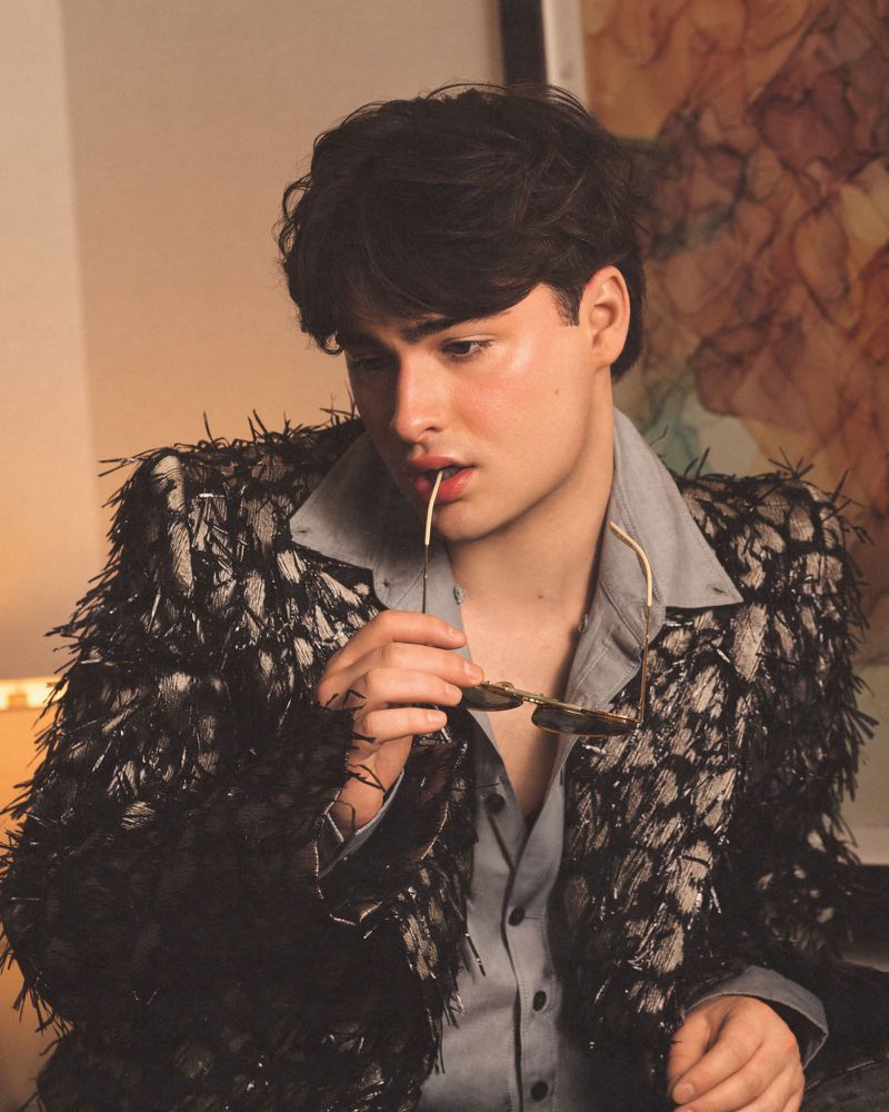 Young man in a textured black and white fringed jacket holding sunglasses to his lips for 1883 Magazine