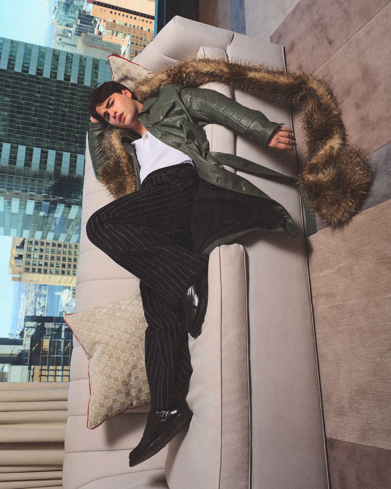 Stylish adult male lounges on a beige couch in pinstripe pants and a green trench coat with a faux fur collar, against a city skyline.