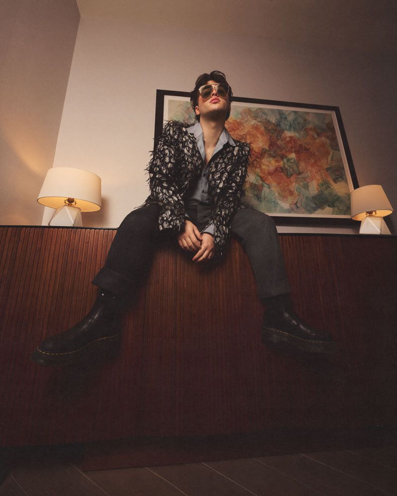 Low-angle shot of a young man seated on a dark wooden cabinet wearing sunglasses, patterned jacket, shirt, trousers, and boots.