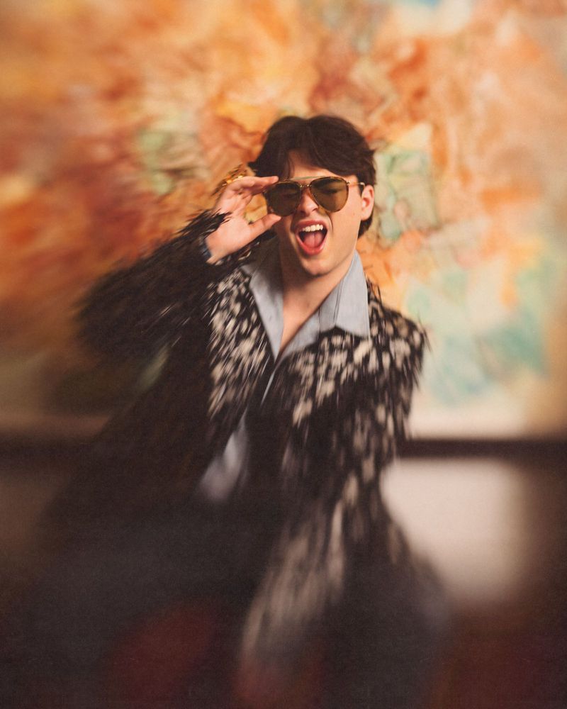 Young man in patterned jacket and sunglasses with motion blur posing in front of abstract art for 1883 Magazine.