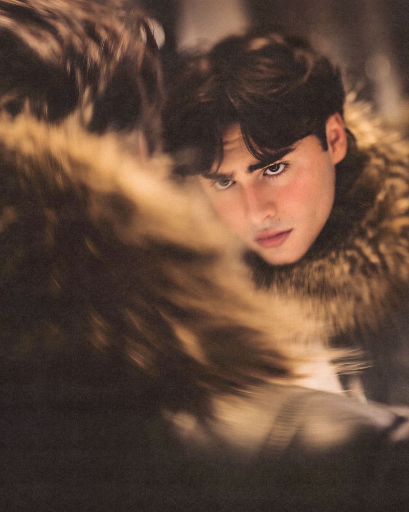 Close-up portrait of a young man with dark hair wearing a furry collar reflected in a mirror captured for 1883 Magazine.