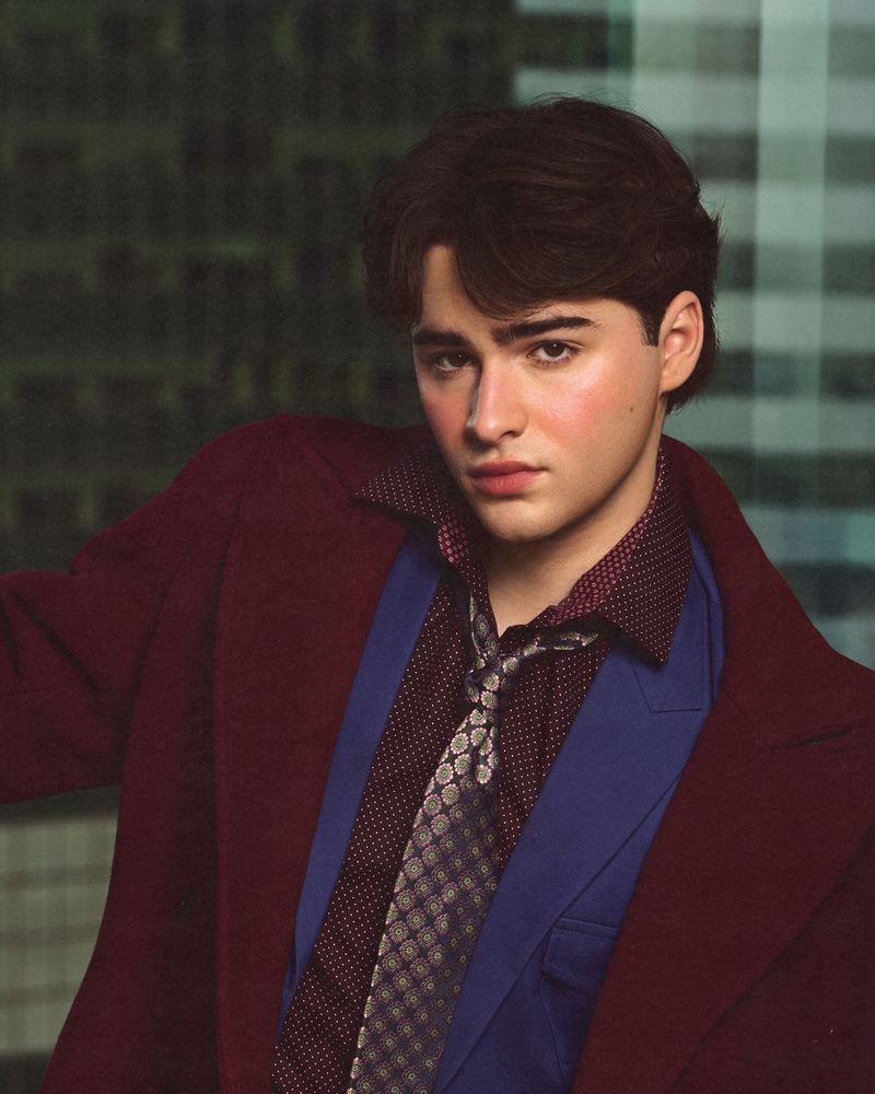 Portrait of young man wearing a burgundy coat, blue blazer, patterned shirt, and tie against a building exterior background