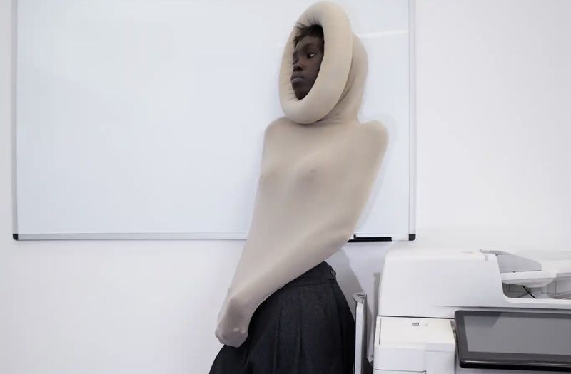 Model wearing a sculptural beige hooded garment and dark skirt leaning against a white wall near a printer.