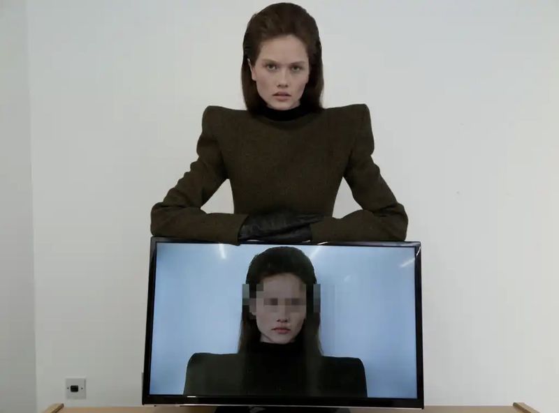 Model in structured brown top and black gloves posing behind a monitor displaying a censored portrait