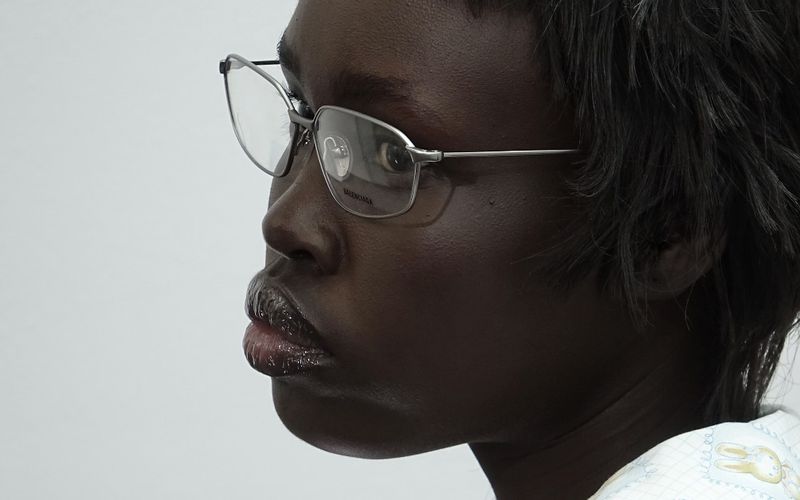 Close-up profile of a dark-skinned adult female wearing silver-rimmed glasses, glossy lip makeup, and a patterned white top against a plain background.
