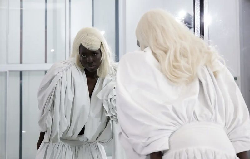 Black woman with long white hair wearing a white gathered blouse looking into a mirror for novembre global