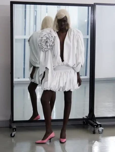 Black model with white hair wearing a white dress with a large flower detail and pink heels standing before a rolling mirror