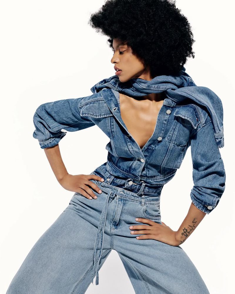 Model with dark curly hair wearing a denim jacket and jeans posing against a white background