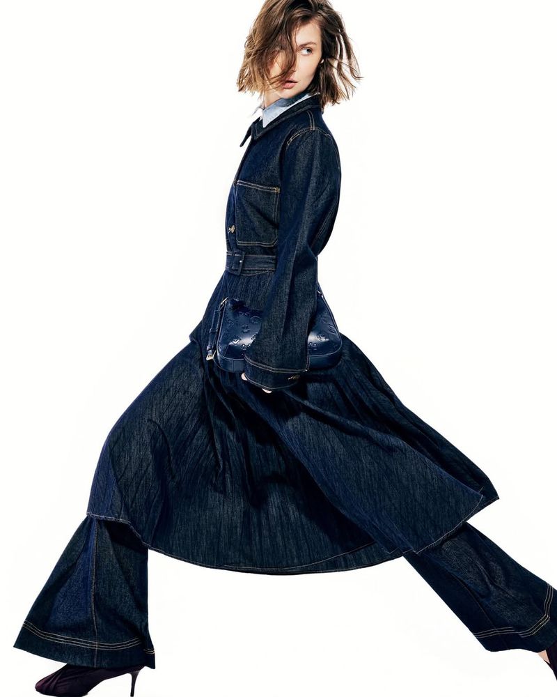Model in a dark denim belted coat and flared trousers holding a blue handbag against a white background.