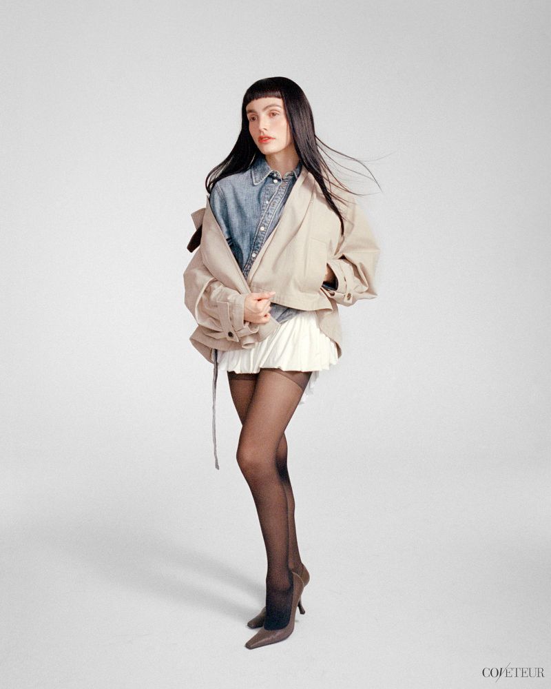 Lux Pascal posing in an oversized beige jacket, denim shirt, white skirt, and black tights against a seamless background.