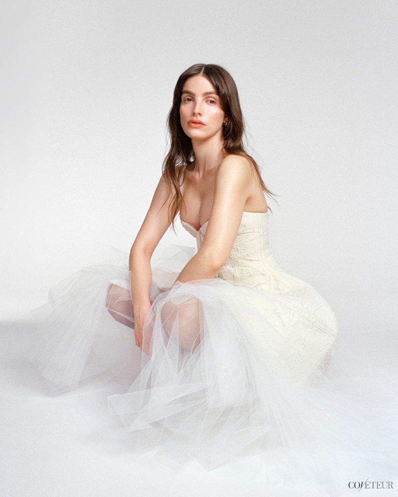 A woman with long brown hair is wearing a cream-colored dress with a tulle skirt in a studio setting with white background.