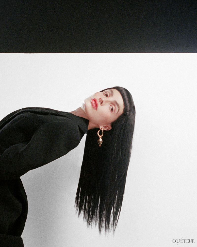 Lux Pascal wearing a black jacket and gold earrings with long dark hair against a white background for Coveteur