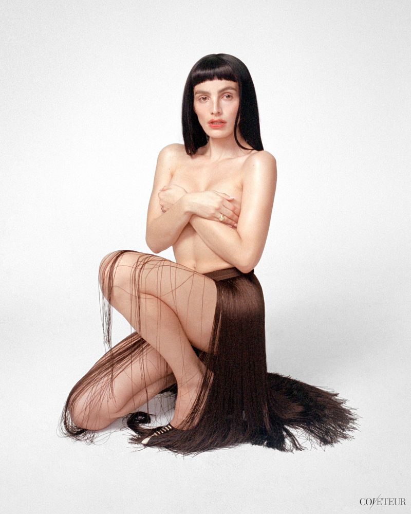 Lux Pascal kneeling on a white seamless backdrop wearing a long fringe skirt and holding hands across chest
