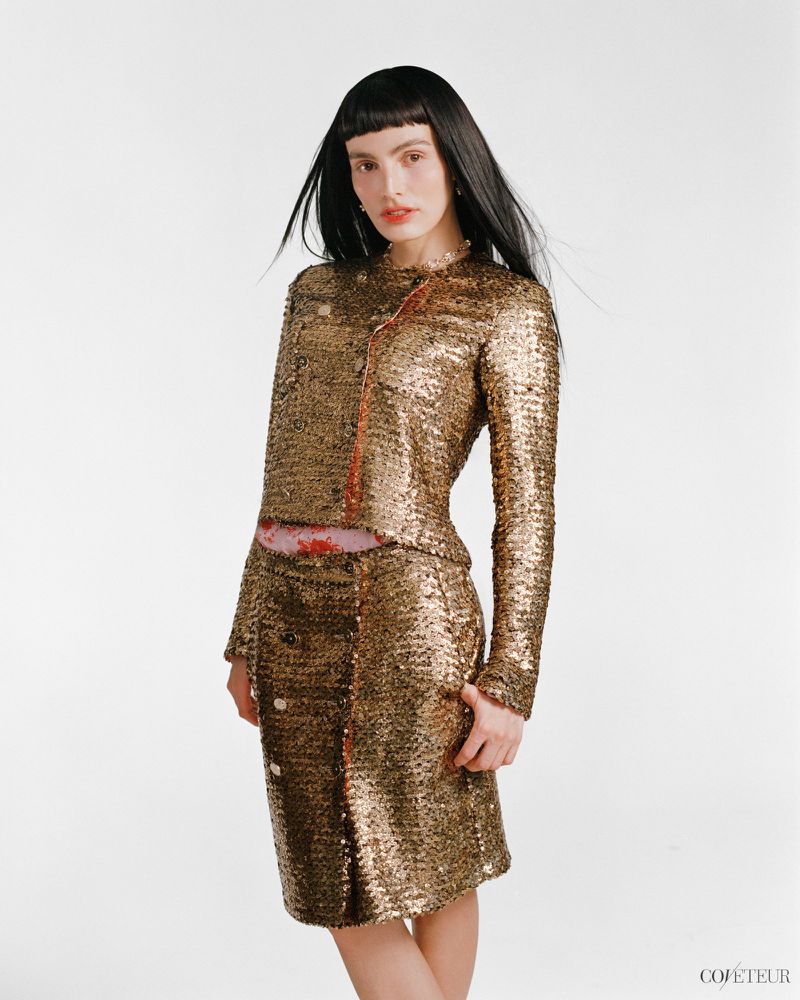 Lux Pascal wearing a gold sequined jacket and matching skirt against a plain white studio background.