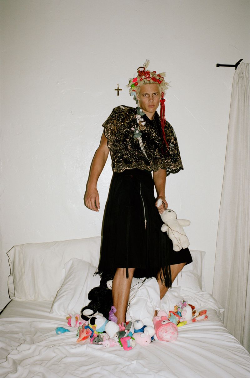 Model Blue River Wild wearing ornate black poncho and skirt standing on bed with plush toys for Tentations Rebelle