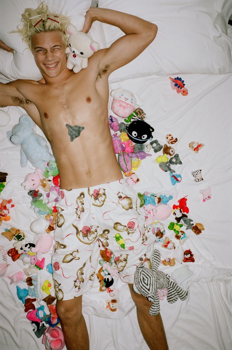 Shirtless male model lying on a white bed surrounded by numerous plush toys and stuffed animals