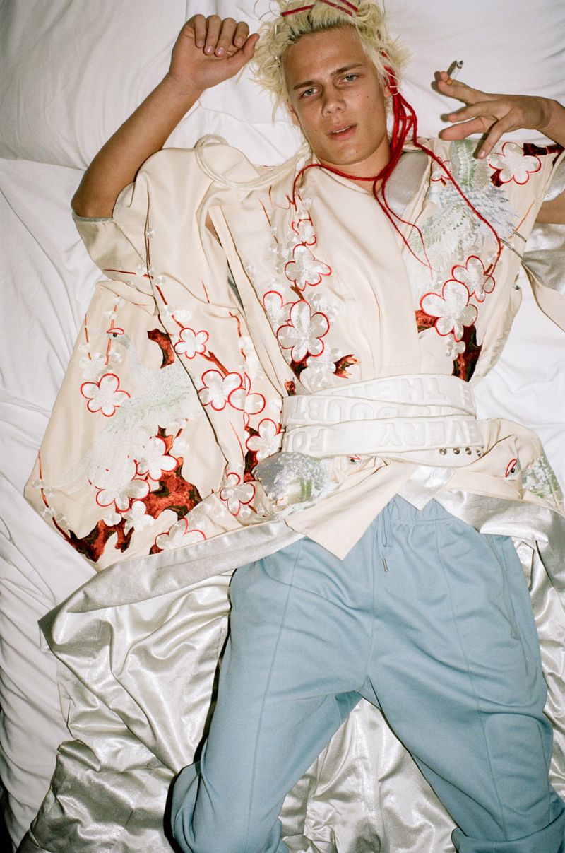 Young adult male lying on a bed with white sheets, wearing a kimono-style garment with floral patterns and light-blue pants while holding a cigarette.