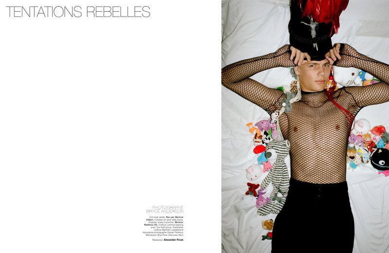 Model in black mesh top and dark trousers lying on a white bed surrounded by small plush toys