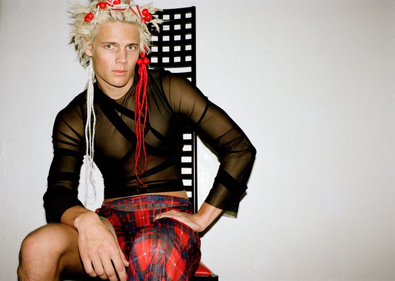 Model with dyed blonde hair wearing a black mesh top and red plaid trousers seated against a white wall.